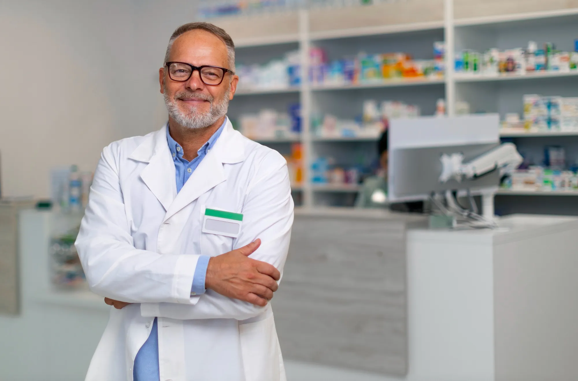 Smiling pharmacist in a white coat with a name badge, arms crossed in a modern pharmacy.